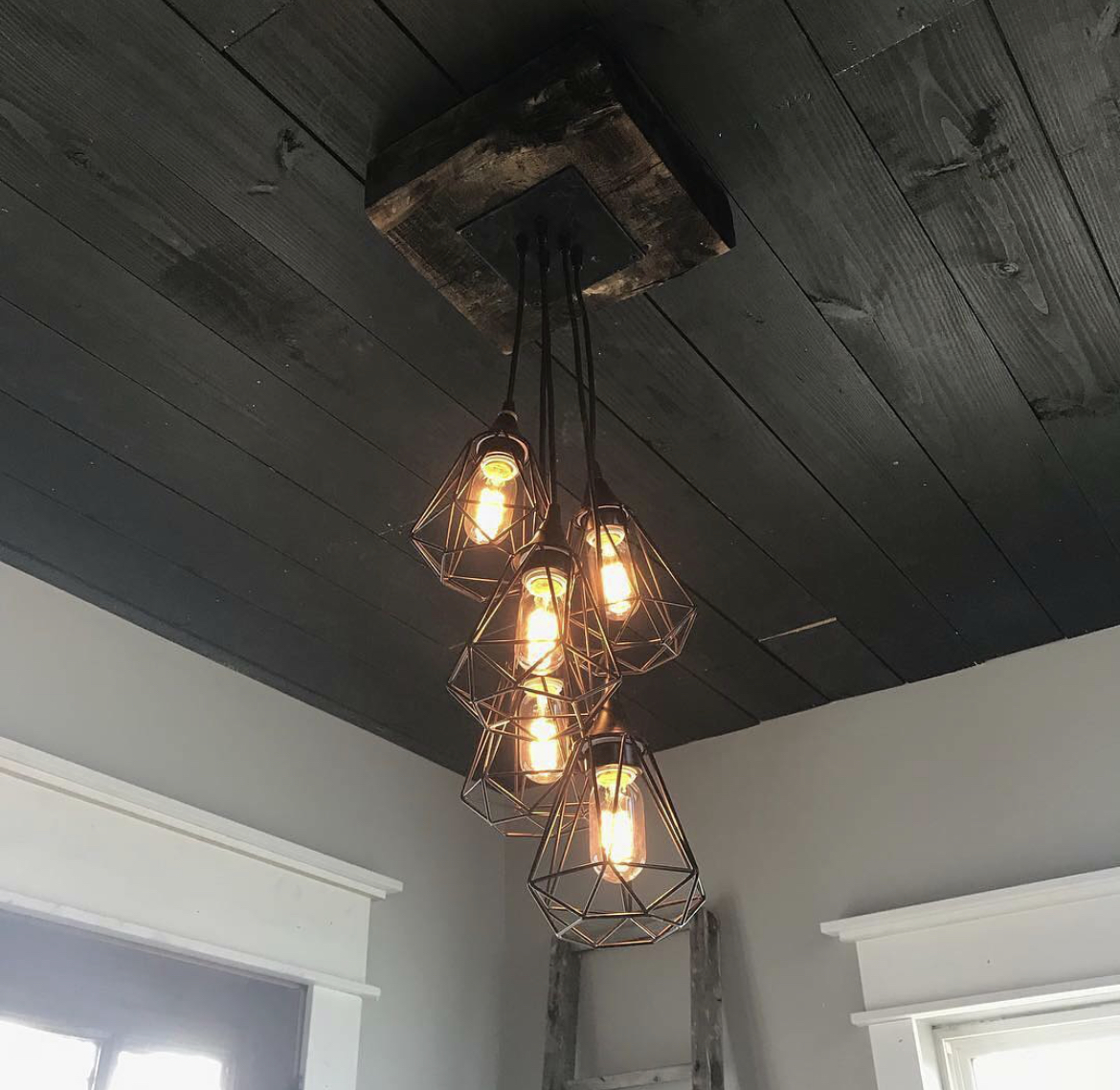 Custom geometric wire cage pendant light cluster with Edison bulbs hanging from reclaimed wood beam on dark-stained ceiling